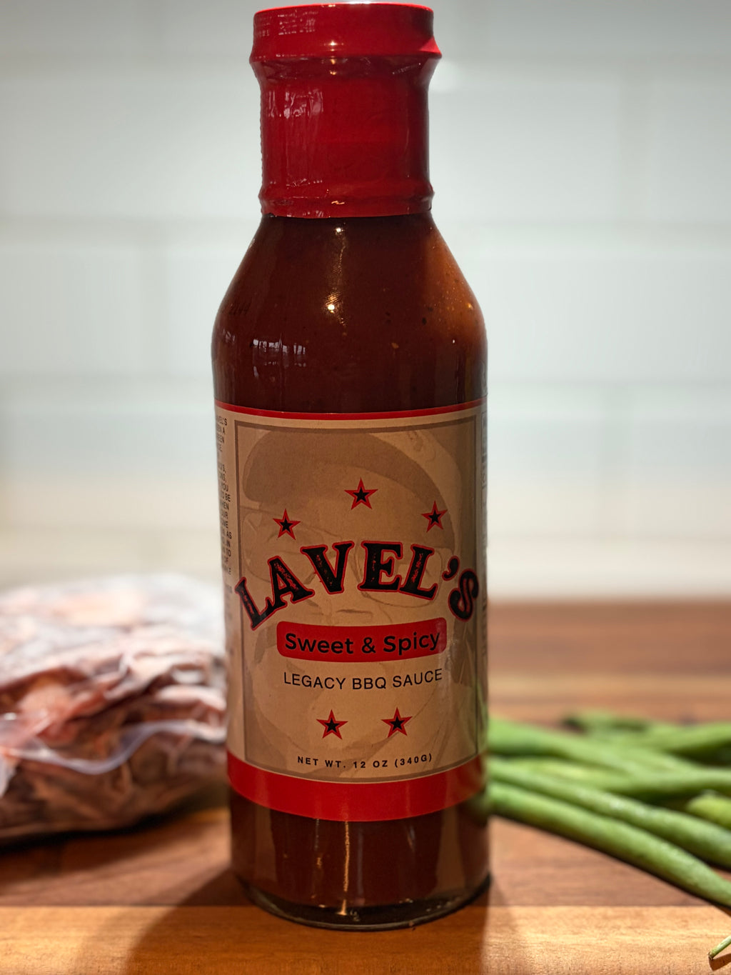 A bottle of Lavel's Sweet & Spicy Legacy BBQ Sauce standing on a wooden cutting board with green beans and packaged meat blurred in the background.